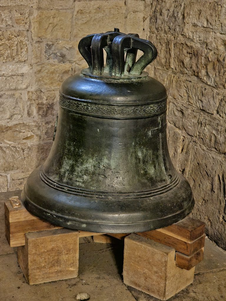 A large, dark-colored bronze or iron bell resting on wooden blocks. The bell has a crown-like structure at the top and a cross emblem on its side. Captured from St. Vitus Cathedral at Prague Castle.