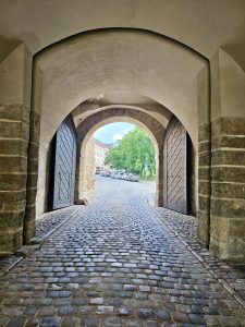 An arched stone gateway with heavy wooden doors, leading to a cobblestone street outside. The gateway is part of historic Vyšehrad Castle in Prague. Through the archway, a glimpse of a street with parked cars and greenery can be seen.