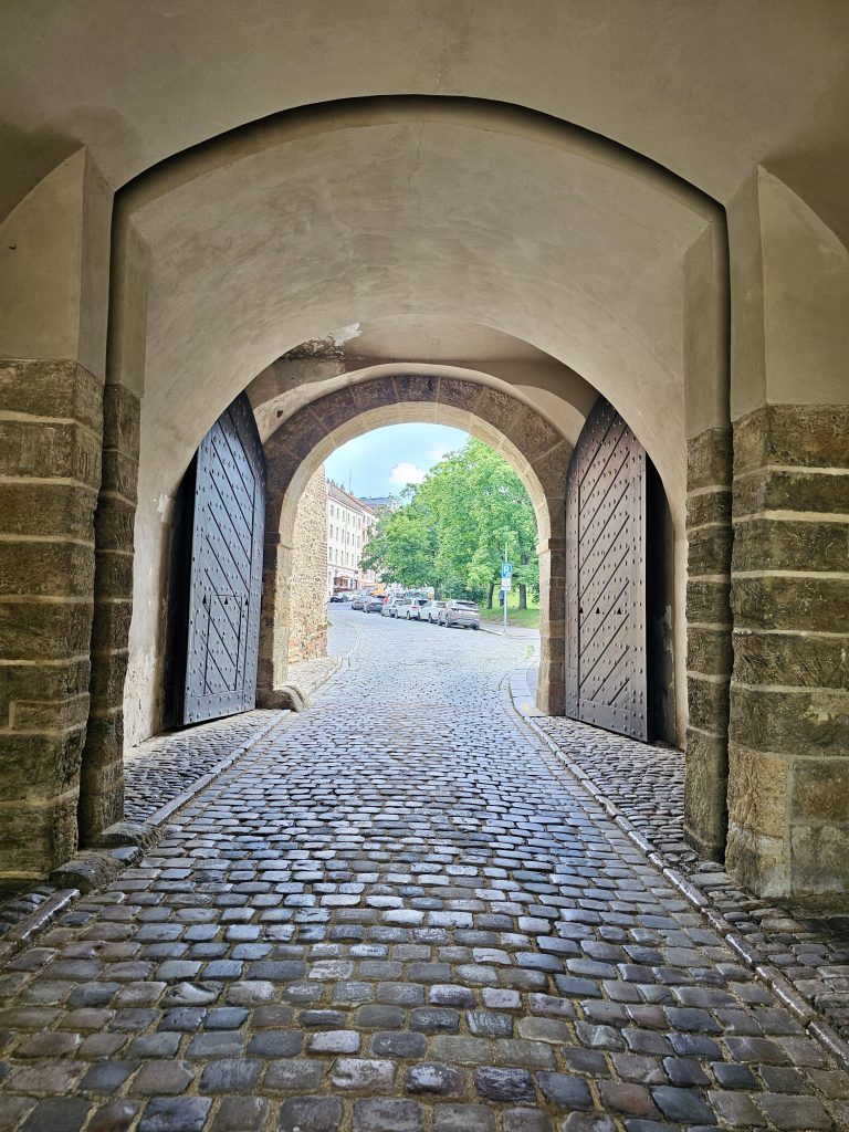 An arched stone gateway with heavy wooden doors, leading to a cobblestone street outside. The gateway is part of historic Vy?ehrad Castle in Prague. Through the archway, a glimpse of a street with parked cars and greenery can be seen.