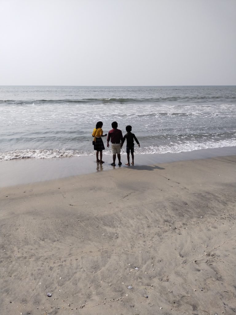 Three children standing on a beach, facing the ocean. The sky is overcast, and gentle waves are approaching the shore.