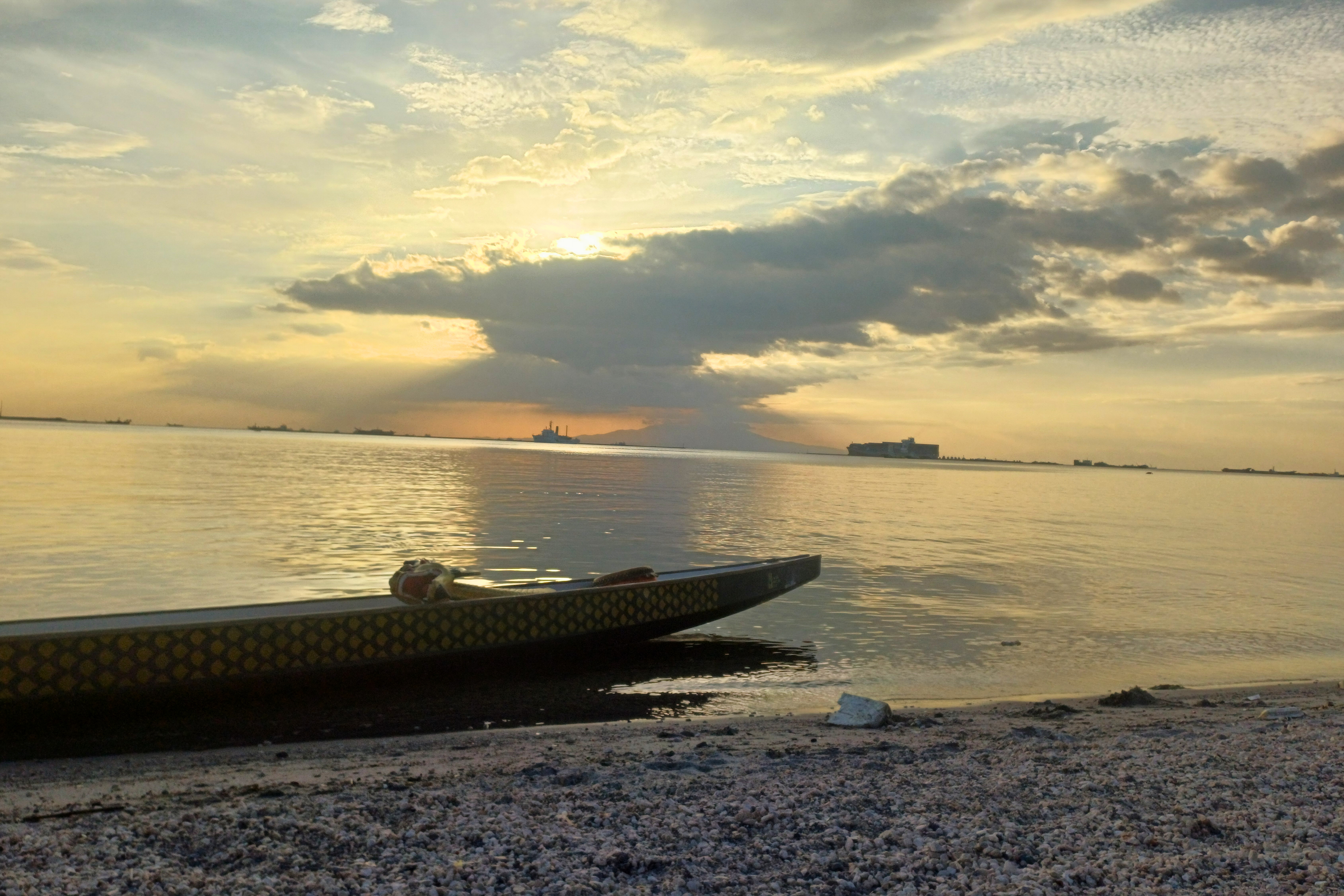 A serene beach scene at sunset, with a long boat resting on the shoreline and calm waters reflecting the golden light of the setting sun. Dark clouds partially cover the sun, casting a dramatic silhouette over the distant horizon. Manila Baywalk