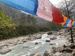 A river flowing through a rocky landscape with green trees on the banks. Colorful prayer flags with inscriptions are strung across the scene, adding vibrancy to the natural setting. Hills covered in dense vegetation are visible in the background.