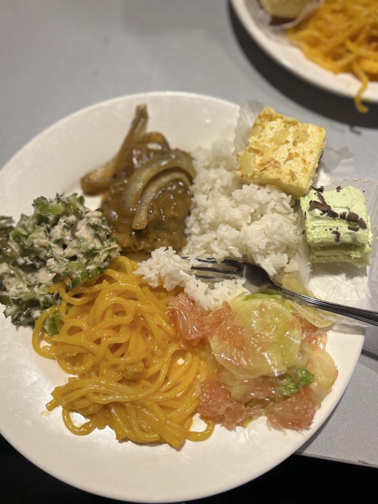 A plate with various Pilipino foods including white rice, noodles, a leafy green salad with a creamy dressing, sautéed onions with meat, a pomelo and cucumber salad, and two small dessert squares, one light yellow and the other light green with chocolate shavings.