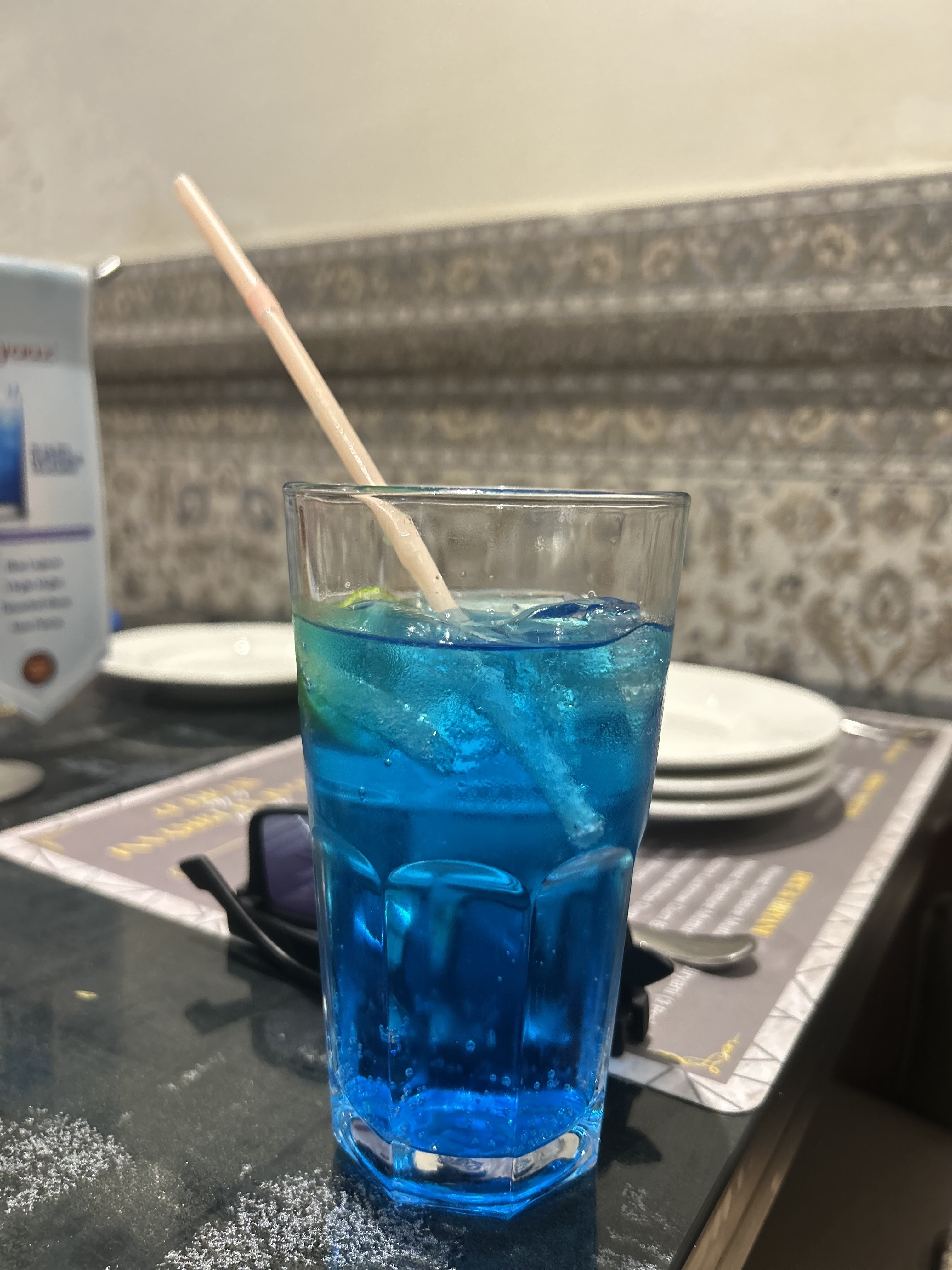 A glass filled with a blue beverage and ice, garnished with a lemon slice, and a straw. It is placed on a table with a menu and stacked white plates in the background.