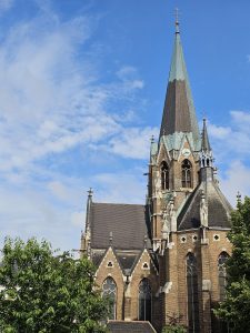 The Lazaristenkirche, a church in Vienna, featuring a tall spire, Gothic architectural details, and a partly cloudy sky backdrop. The church's facade is predominantly brown brick, with darker roofing and some greenery visible at the bottom.