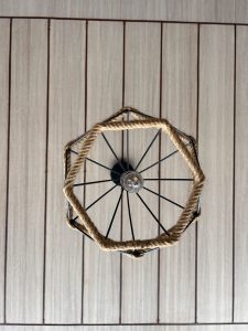 A ceiling light fixture with a central bulb, surrounded by spokes forming a radial pattern, and an octagonal frame wrapped with rope, against a wooden panel ceiling.