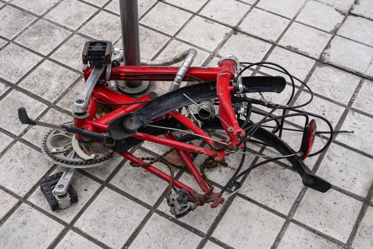 A dismantled and rusted red bicycle frame lying on a tiled ground, secured by a lock to a metal pole. Missing wheels and other parts, with tangled cables visible.