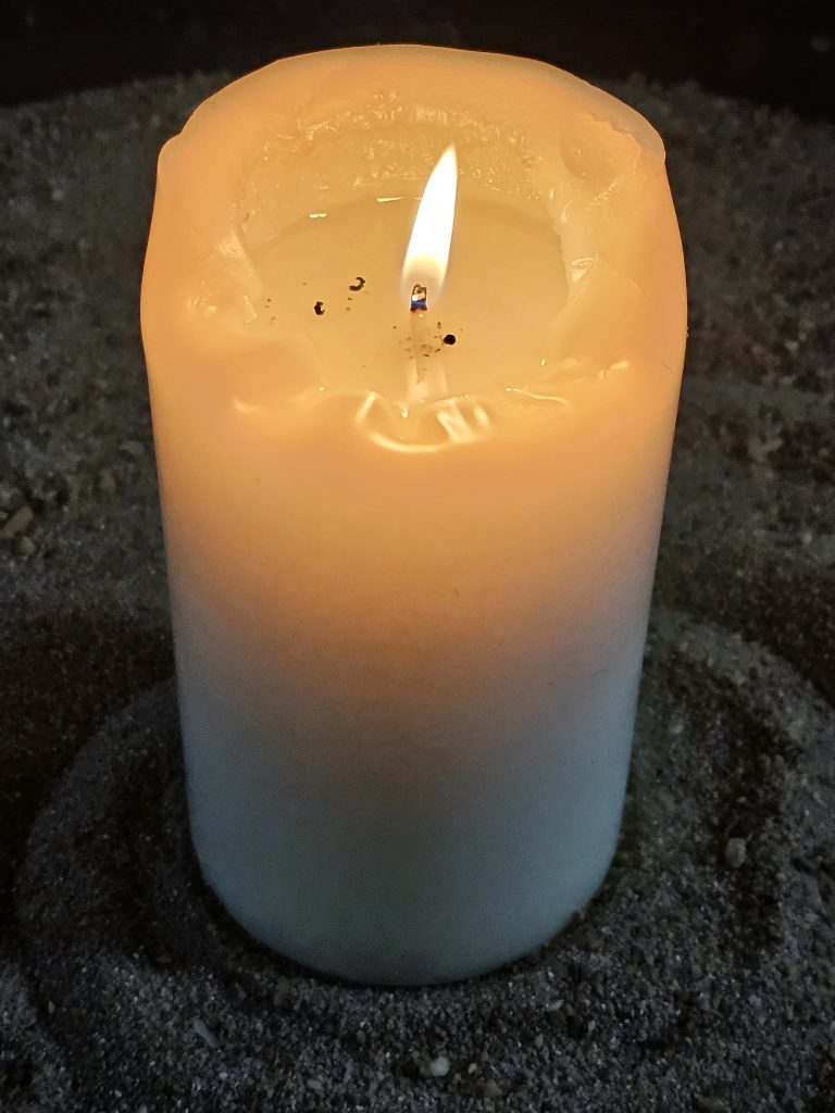 A thick candle burning. The candle is a pale yellow color, fading to a light blue at the bottom, and it sits in a bed of dark grey sand. From  Andr?kirche (St. Andr? Church) in Salzburg.