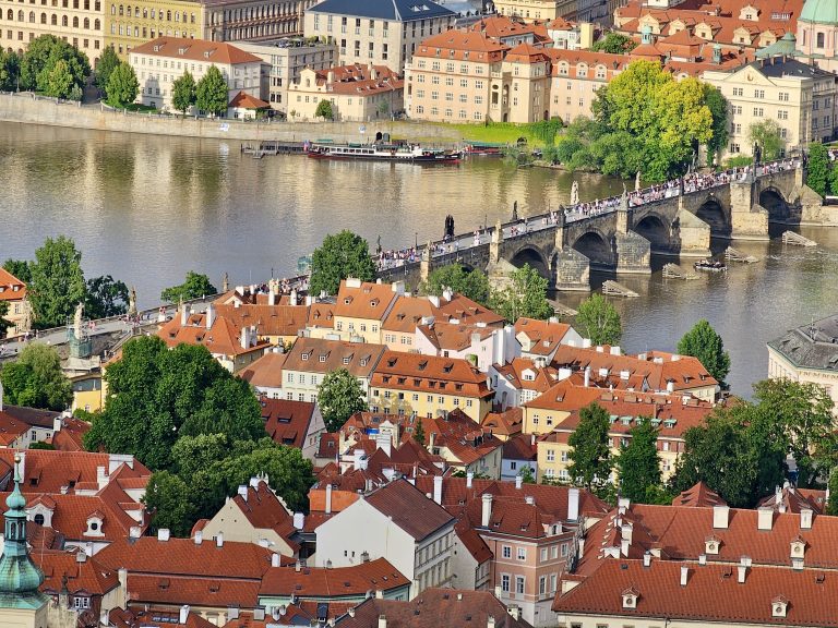 An aerial view of Prague from Petrin Tower featuring the Charles Bridge spanning the Vltava river, with the old town’s red-roofed buildings clustered on either side. The bridge is filled with pedestrians, and the architecture of the buildings suggests a historical European cityscape.