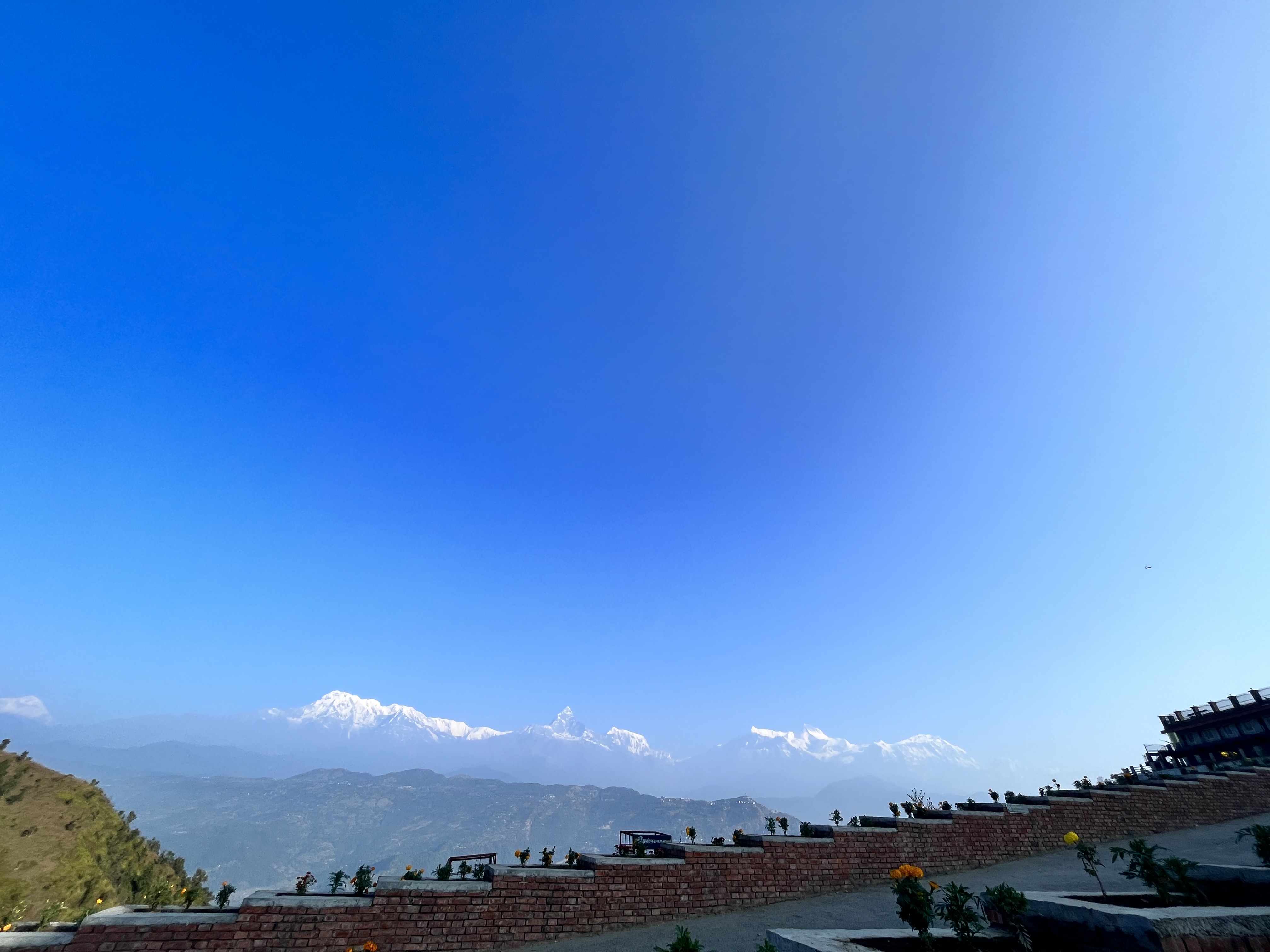 A scenic view of snow-capped mountains under a clear blue sky, with a brick wall and flowers in the foreground.