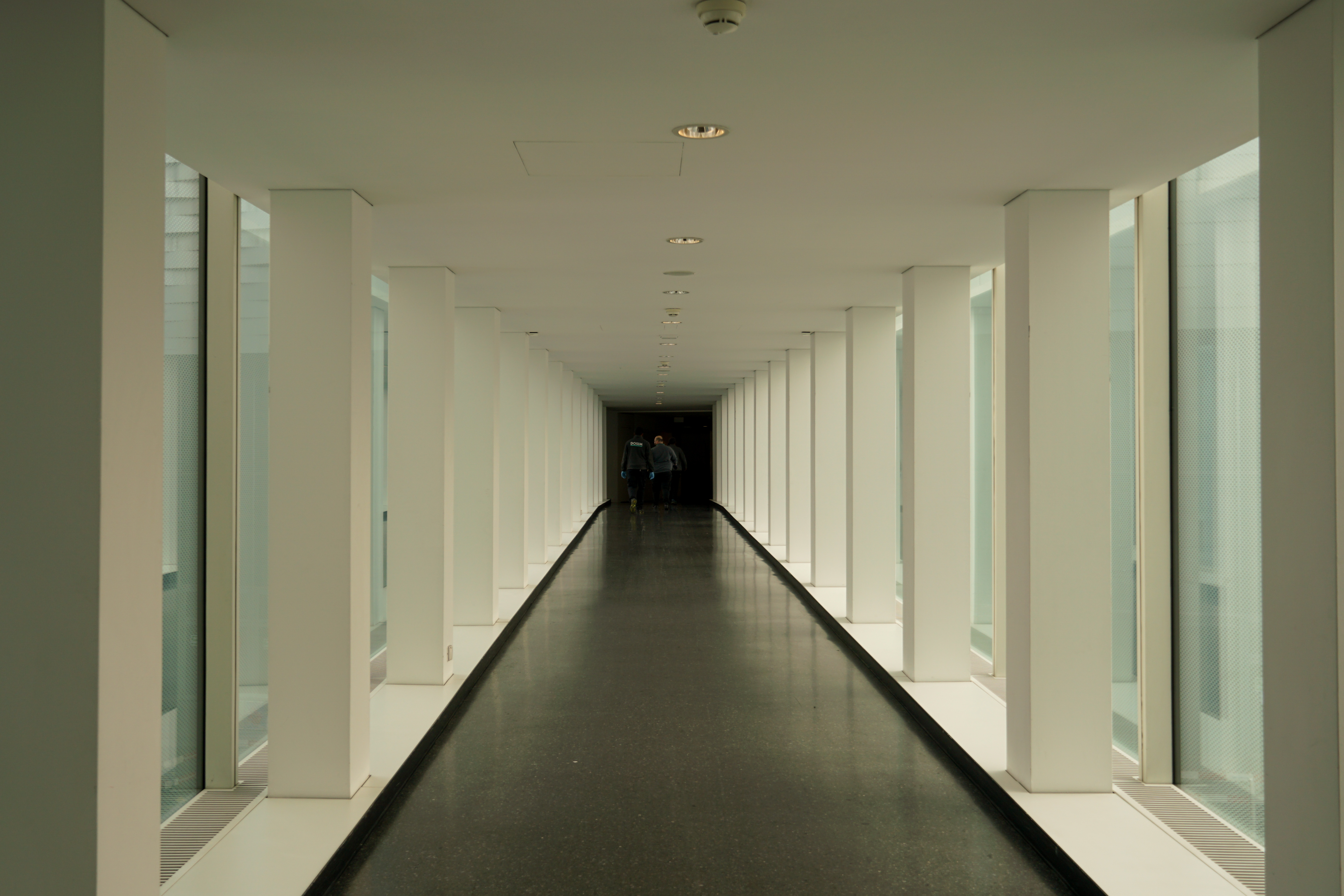 A long, modern corridor with large windows on both sides and two people walking in the distance.