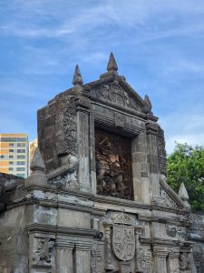 菲律宾马尼拉圣地亚哥堡（Fort Santiago）的尖顶结构是一个古老的石头立面，上面雕刻着华丽的雕刻和历史场景的浮雕。这座建筑显示出风化的迹象。背景是现代建筑和晴朗蓝天下的一棵树。
