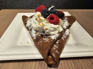 A chocolate crepe folded into a triangular shape, topped with whipped cream, raspberries, blueberries, powdered sugar, and chopped nuts, presented on a white square plate against a wood-like surface. Captured from a restaurant in Turin, Italy.  