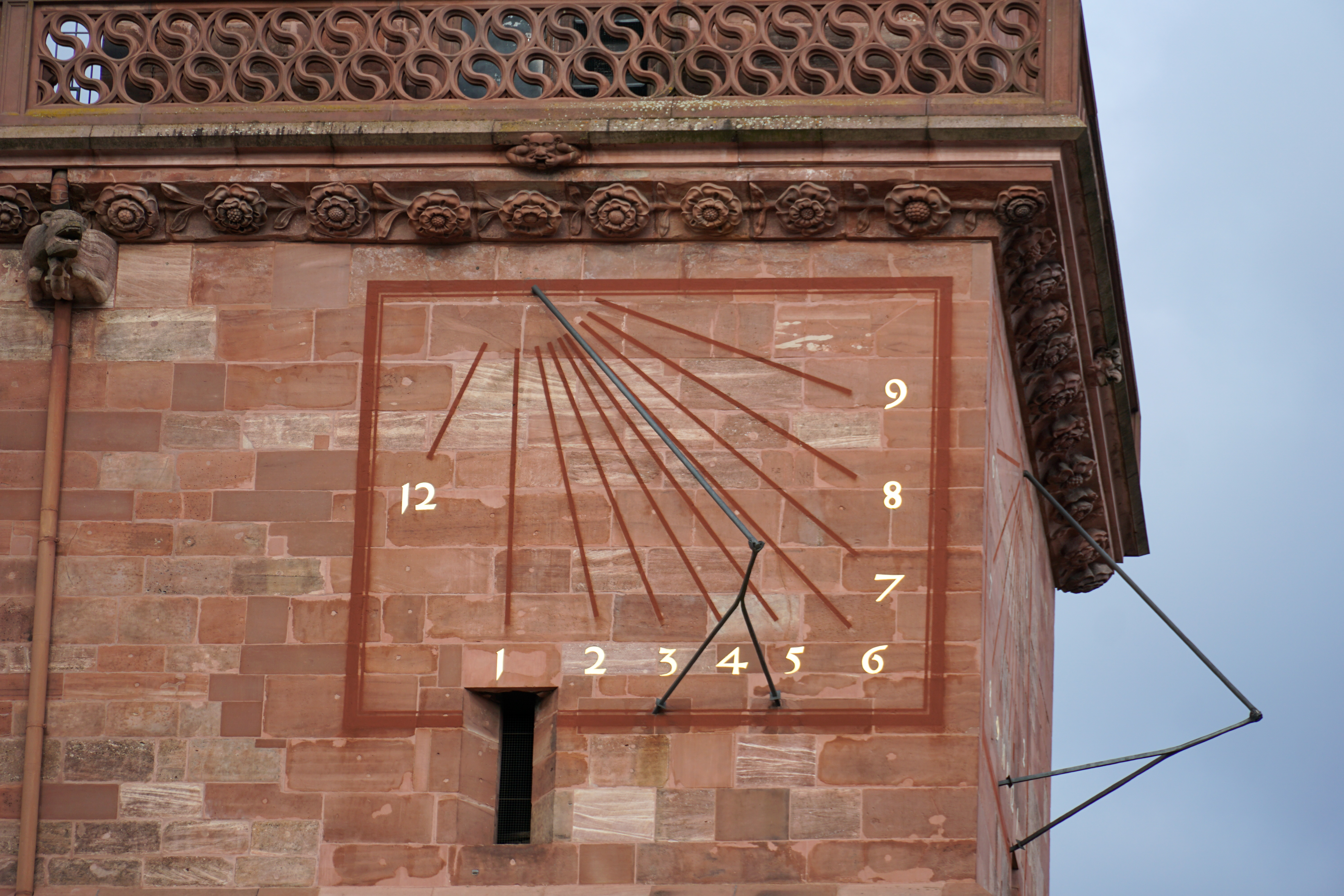 A historic stone building wall features an ornate sundial with a gnomon and painted hour lines, marked with numbers, accompanied by floral carvings and decorative brickwork above.