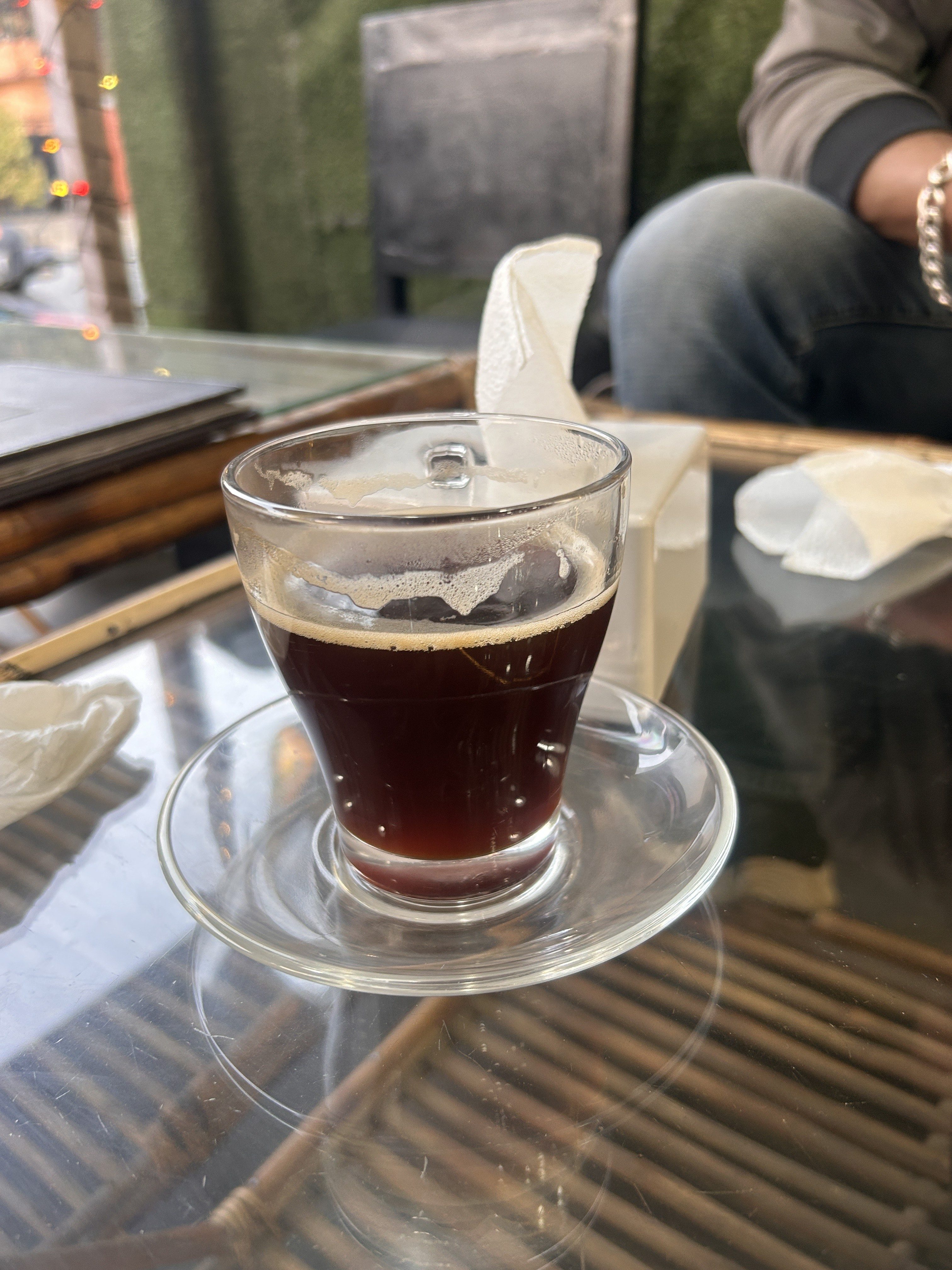 A glass cup filled with dark coffee sits on a clear glass saucer on a glass table.