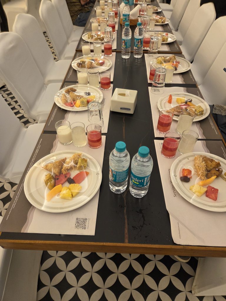 A long table set for a meal, with white chairs on both sides. Each place setting has a white plate filled with a variety of appetizers and fruits, and several glasses containing different colored beverages.