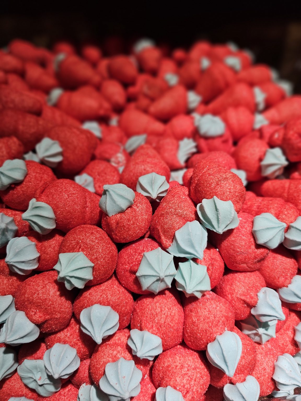 A close-up of many strawberry-shaped marshmallows. The marshmallows are primarily red with a light blue, flower-shaped piece on top, resembling the green leafy part of a strawberry.