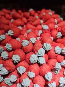 A close-up of many strawberry-shaped marshmallows. The marshmallows are primarily red with a light blue, flower-shaped piece on top, resembling the green leafy part of a strawberry.
