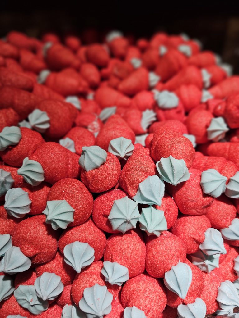 A close-up of many strawberry-shaped marshmallows. The marshmallows are primarily red with a light blue, flower-shaped piece on top, resembling the green leafy part of a strawberry.