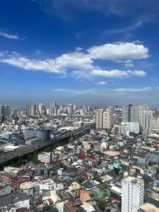 A vibrant aerial view of Manila, Philippines, showcasing a mix of colorful low-rise residential buildings and towering skyscrapers under a bright blue sky with scattered white clouds. 