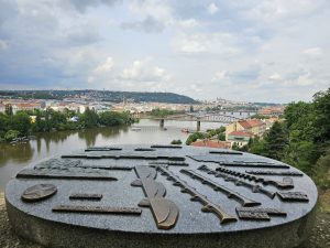 A panoramic view of Prague, taken from Vy?ehrad, with a detailed metal relief map in the foreground. The Vltava River flows through the city, spanned by several bridges, including the Railway Bridge (?elezni?ní most).