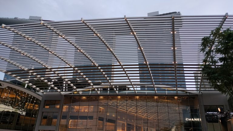 Modern architectural canopy at The Shoppes at Marina Bay Sands, Singapore, with reflections of city lights and towering skyscrapers in the background.