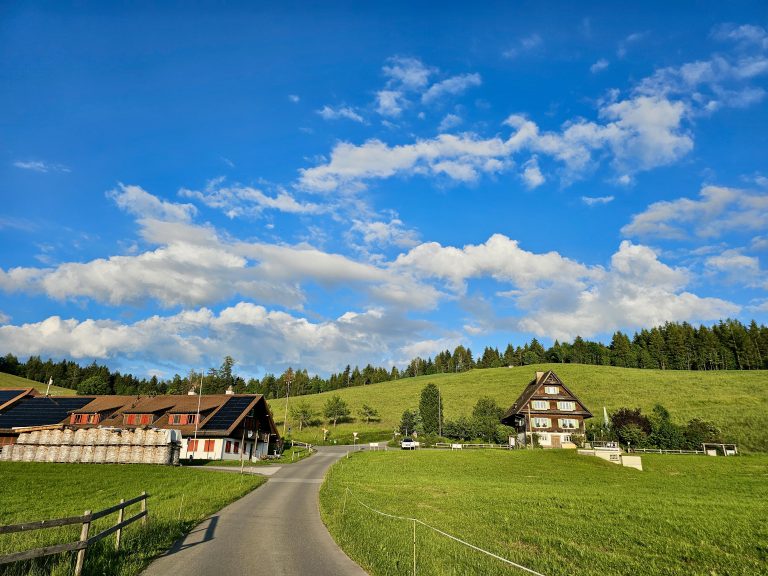 A landscape in Zugerberg, Swiss featuring green fields, two houses, a road, a forest in the background, and a blue sky with white clouds.