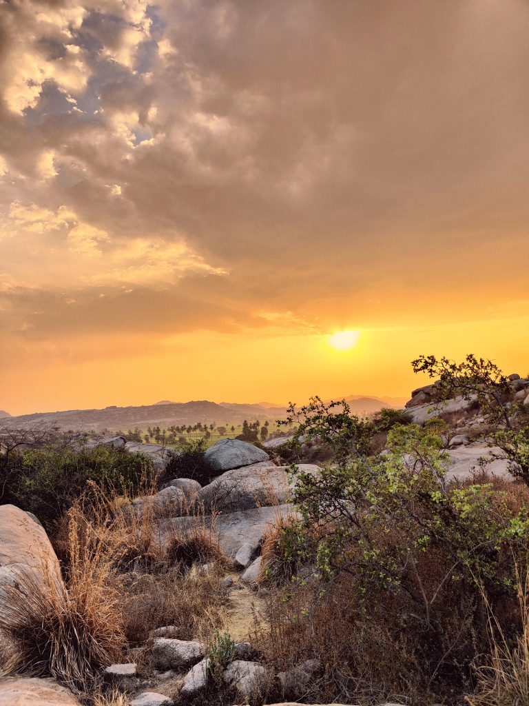 A scenic landscape of a rocky desert area at sunset. The sky is filled with warm hues of orange and yellow, interspersed with clouds. The foreground features rocks, dry grasses, and some green shrubs. The background shows distant mountains and a line of palm trees, all under the vivid glow of the setting sun. Photo taken at Hampi, India.