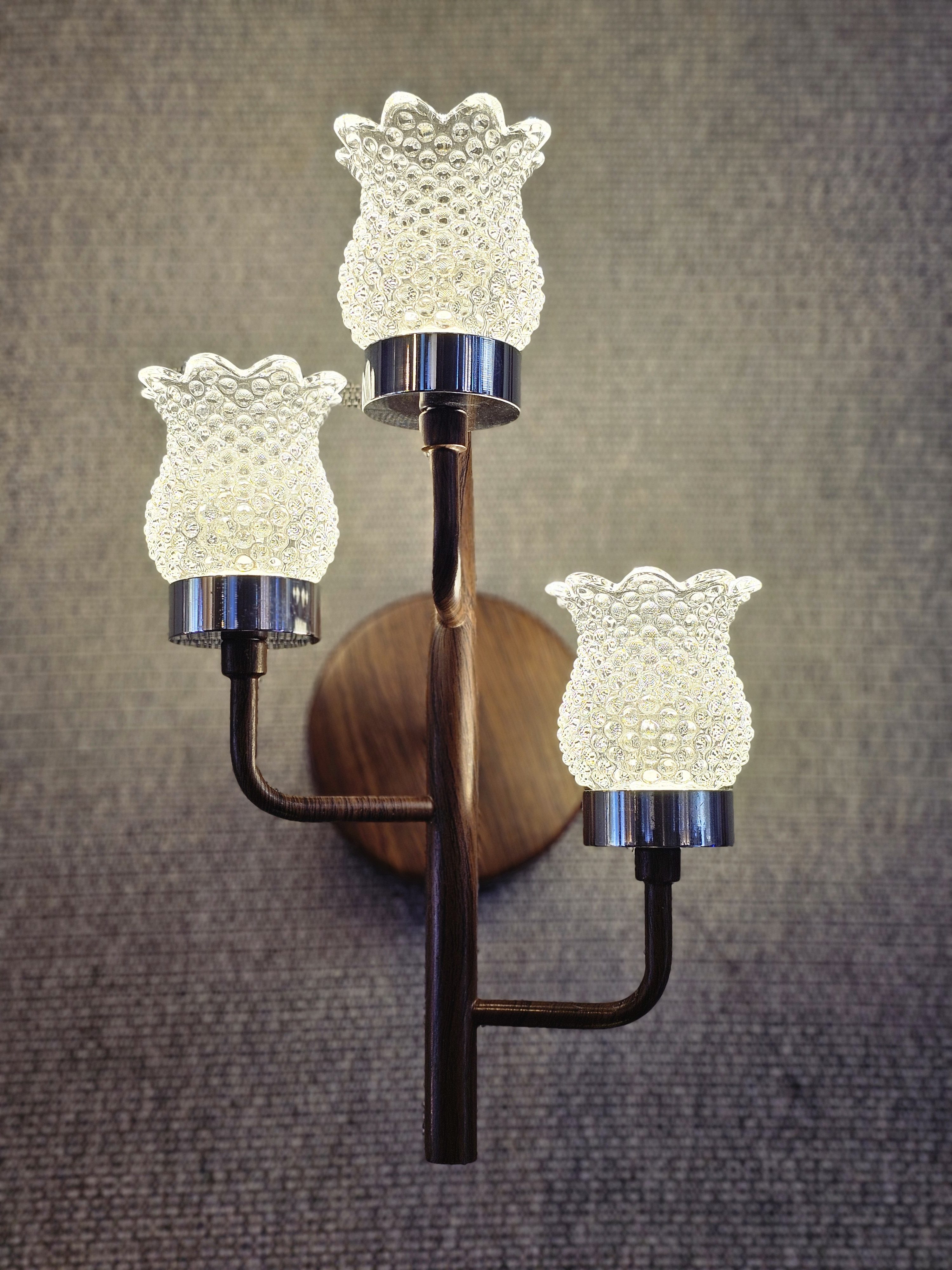 Three glowing glass shades are mounted on the wall. Each shade has a scalloped edge and textured surface. The light fixture blends wood and metal for a unique design. Captured from an electrical shop in Kozhikode.