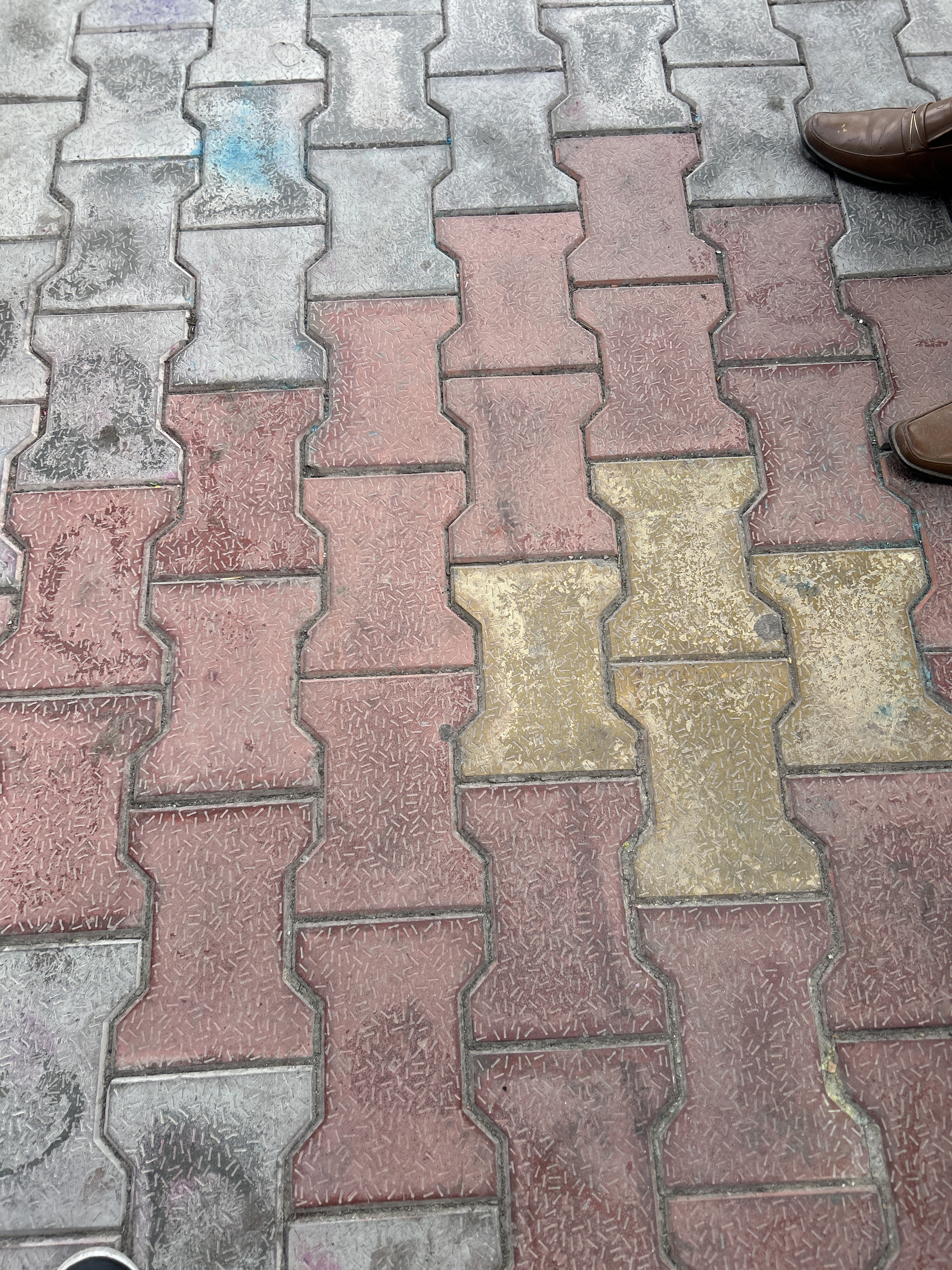 Interlocking colored pavement tiles in shades of gray, red, and yellow. Partially visible brown shoes appear on the right side of the image.