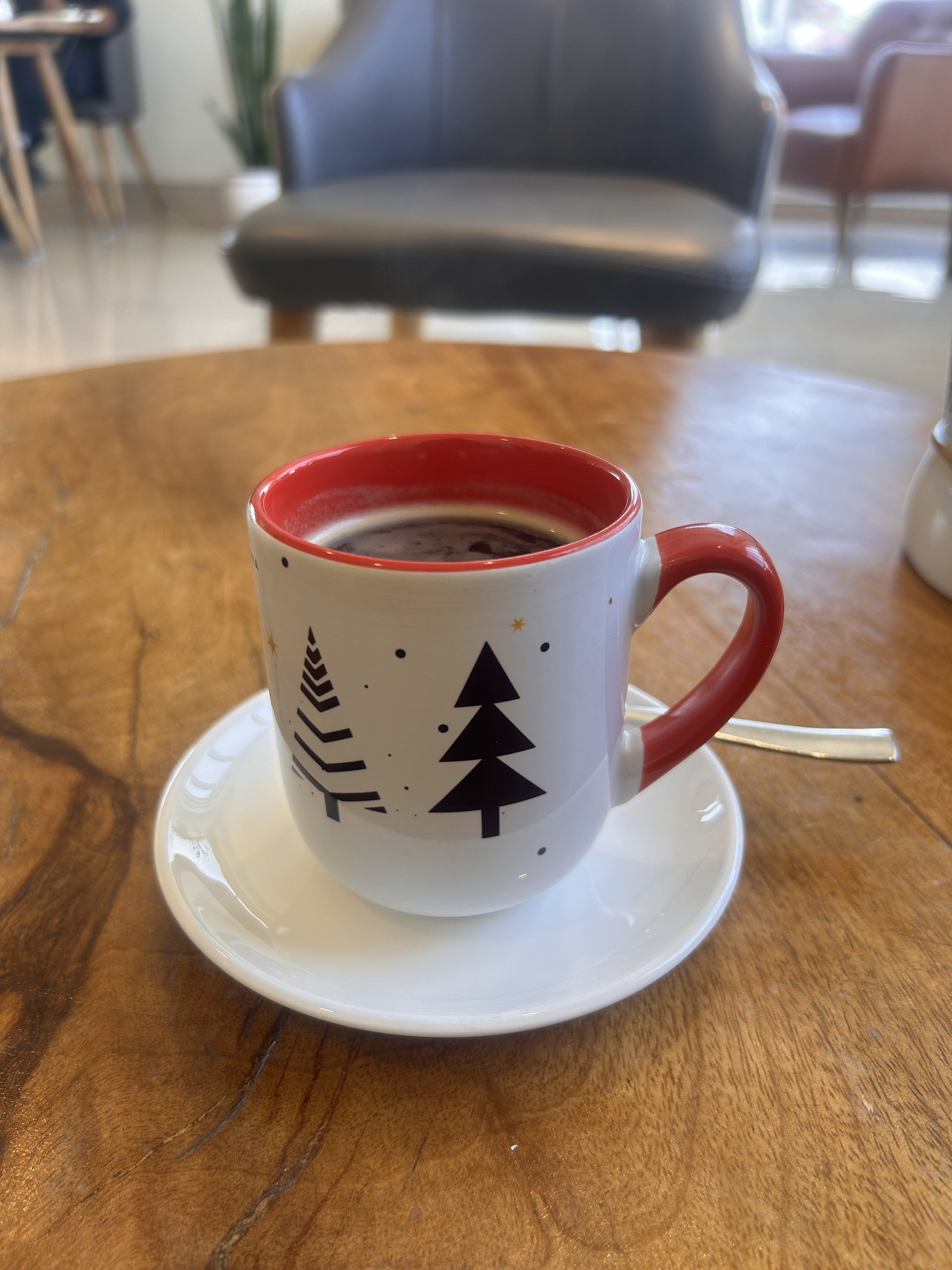 A cup of coffee in a festive mug with a red interior and Christmas tree designs sits on a saucer on a wooden table in a cozy cafe setting.