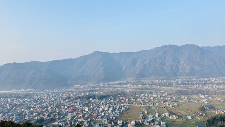 Aerial view of a cityscape with numerous buildings dispersed across a valley, surrounded by lush green hills and mountains under a clear blue sky.