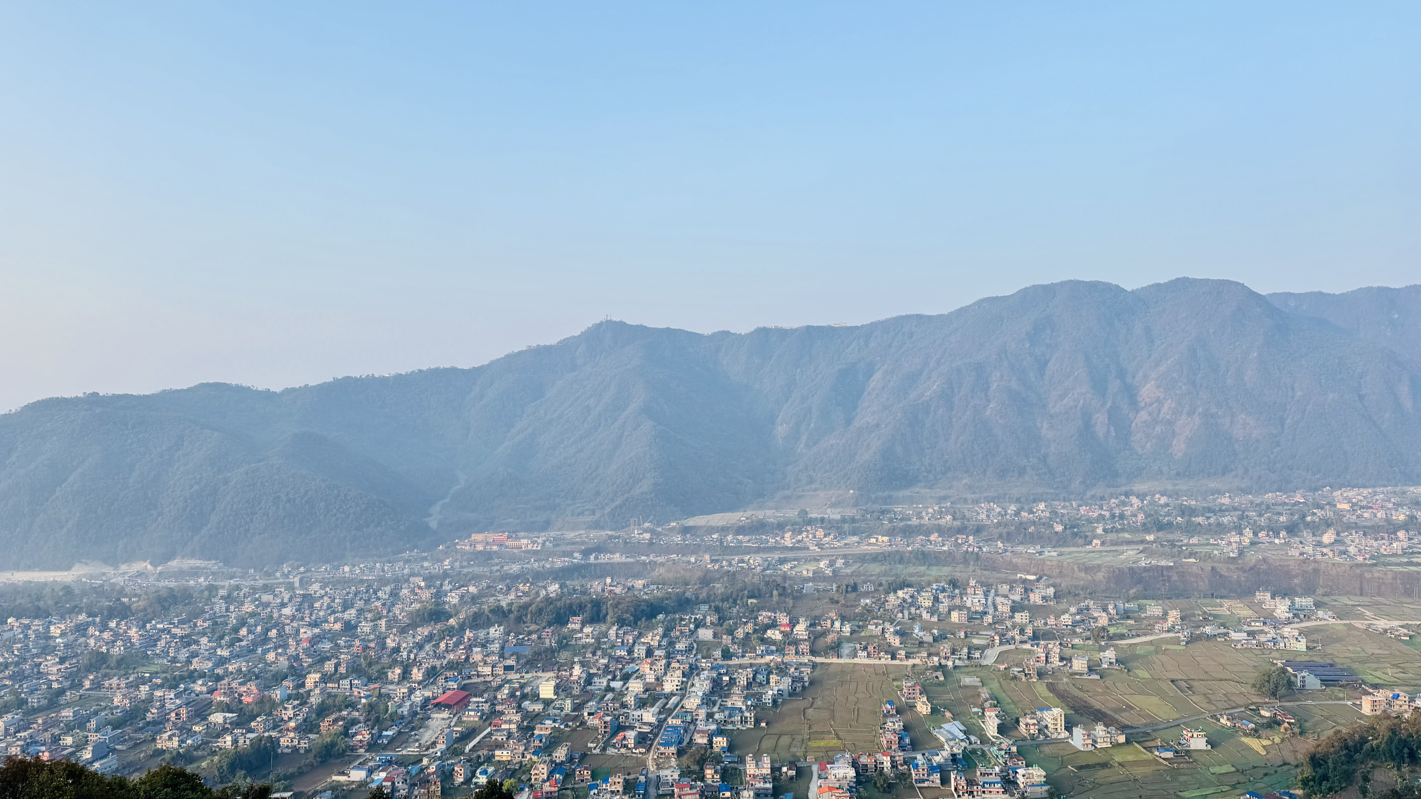 Aerial view of a cityscape with numerous buildings dispersed across a valley, surrounded by lush green hills and mountains under a clear blue sky.
