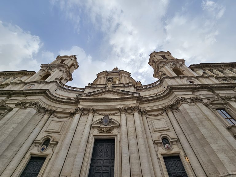 A low angle view of the facade of Sant’Agnese in Agone, a 17th-century Baroque church in Rome, showcasing its ornate architectural details. The pale stone structure emphasizes the height and grandeur of the building. The composition highlights the church’s towers, dome, and the intricate carvings above its entrance.