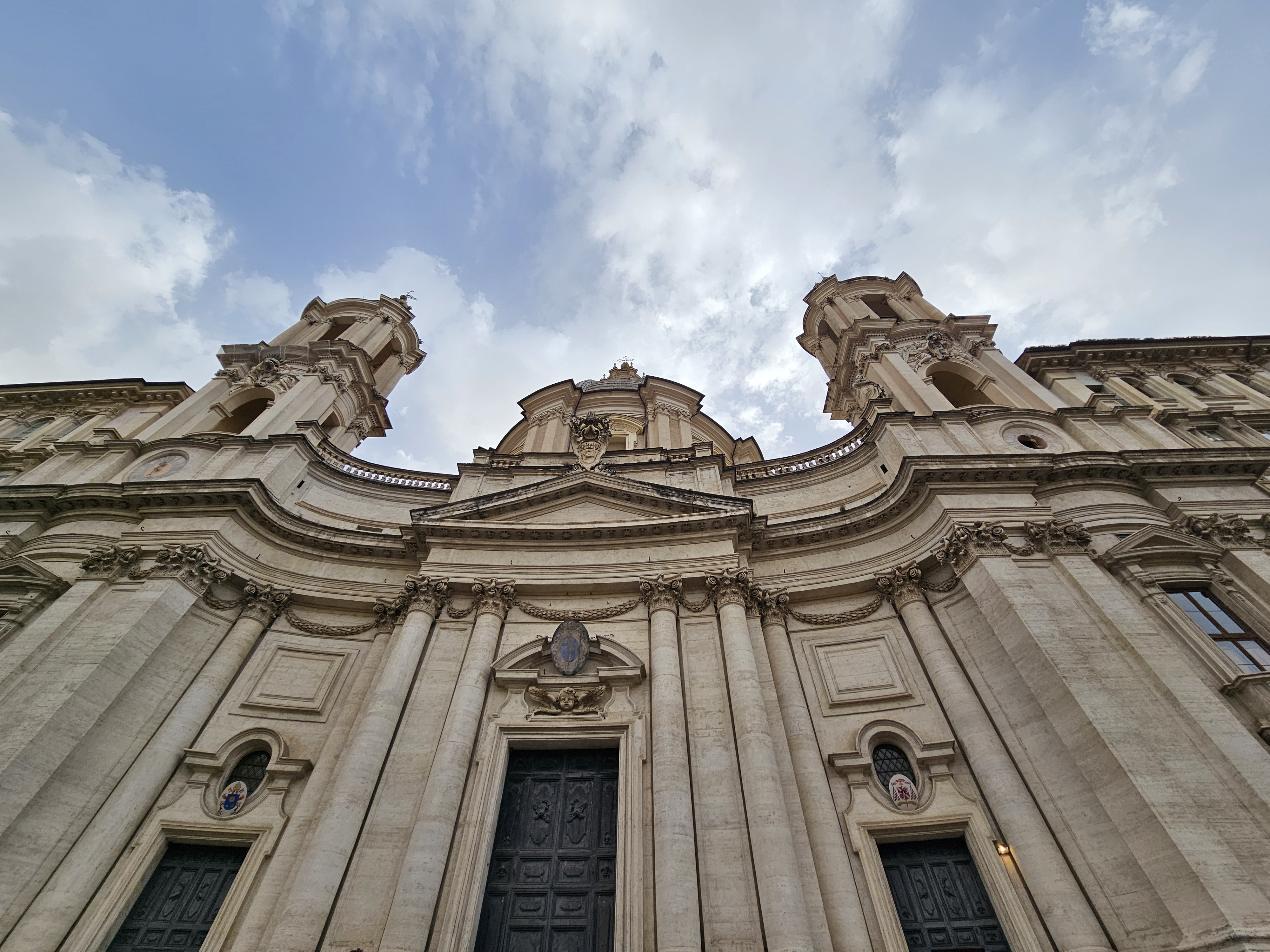A low angle view of the facade of Sant'Agnese in Agone, a 17th-century Baroque church in Rome, showcasing its ornate architectural details. The pale stone structure emphasizes the height and grandeur of the building. The composition highlights the church's towers, dome, and the intricate carvings above its entrance.
