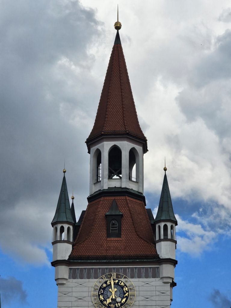 The upper portion of the Old Town Hall Clock Tower in Munich, Germany, features its distinctive red-tiled roof, ornate white facade, and a clock face. The tower is set against a backdrop of a cloudy sky, highlighting its architectural details and the contrast between the warm tones of the building and the cool tones of the sky.