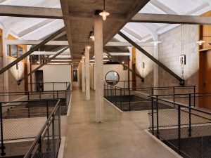 An industrial-style corridor with concrete beams and overhead lighting, featuring metal railings and mesh dividers. The corridor has a cement floor and numbered doors on the right, with a circular window visible in the background.