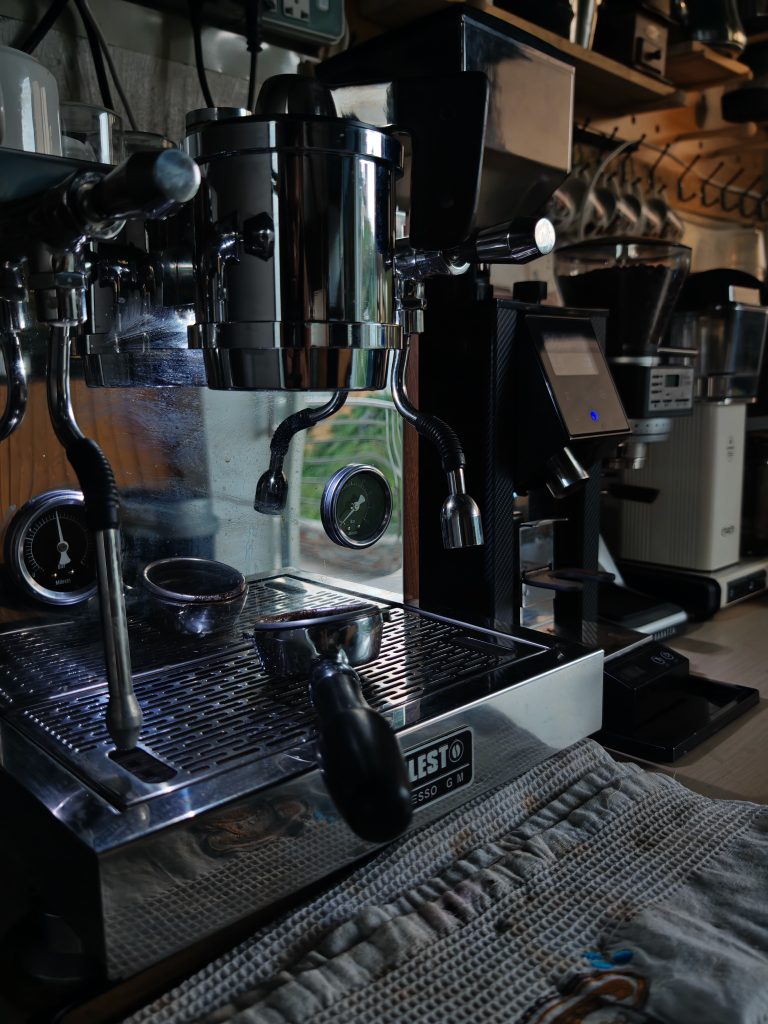 A close-up of a professional espresso machine with a portafilter resting on the drip tray. In the background are shelves with coffee-making equipment and accessories. The setting appears to be a cozy and well-equipped coffee bar.