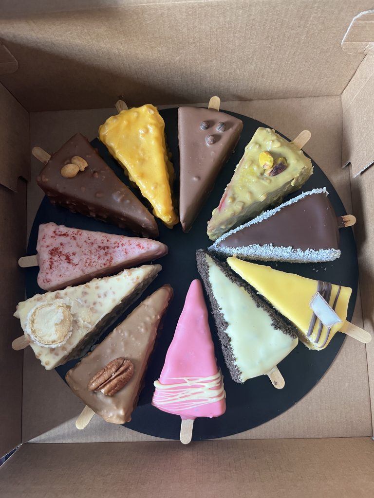 An assortment of colorful, triangular cake slices on sticks arranged in a circular pattern on a black platter. Each slice has different toppings and frosting, creating a variety of textures and colors.