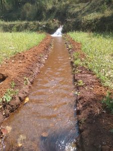 A narrow, man-made water channel runs through a grassy area, with water flowing down a small slope surrounded by earth banks, leading into a lush, green landscape.