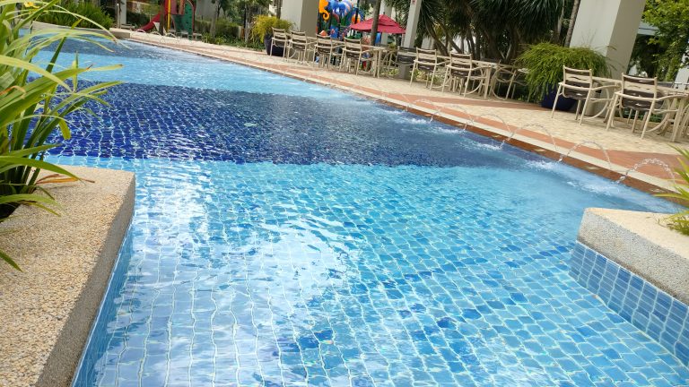 A clear swimming pool with blue mosaic tiles and water jets creating arcs across the surface. Surrounding the pool are chairs and tables in a patio area, with lush greenery and a playground in the background.