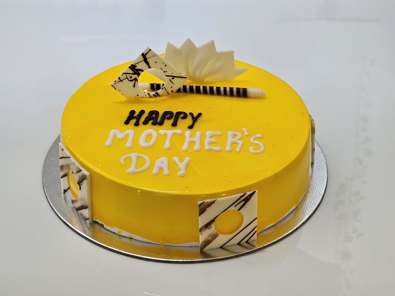 A round Mango cake with bright yellow frosting, decorated with white chocolate pieces and a white fondant flower. The cake has “HAPPY MOTHER’S DAY” written on it in white lettering and is placed on a silver cake board.