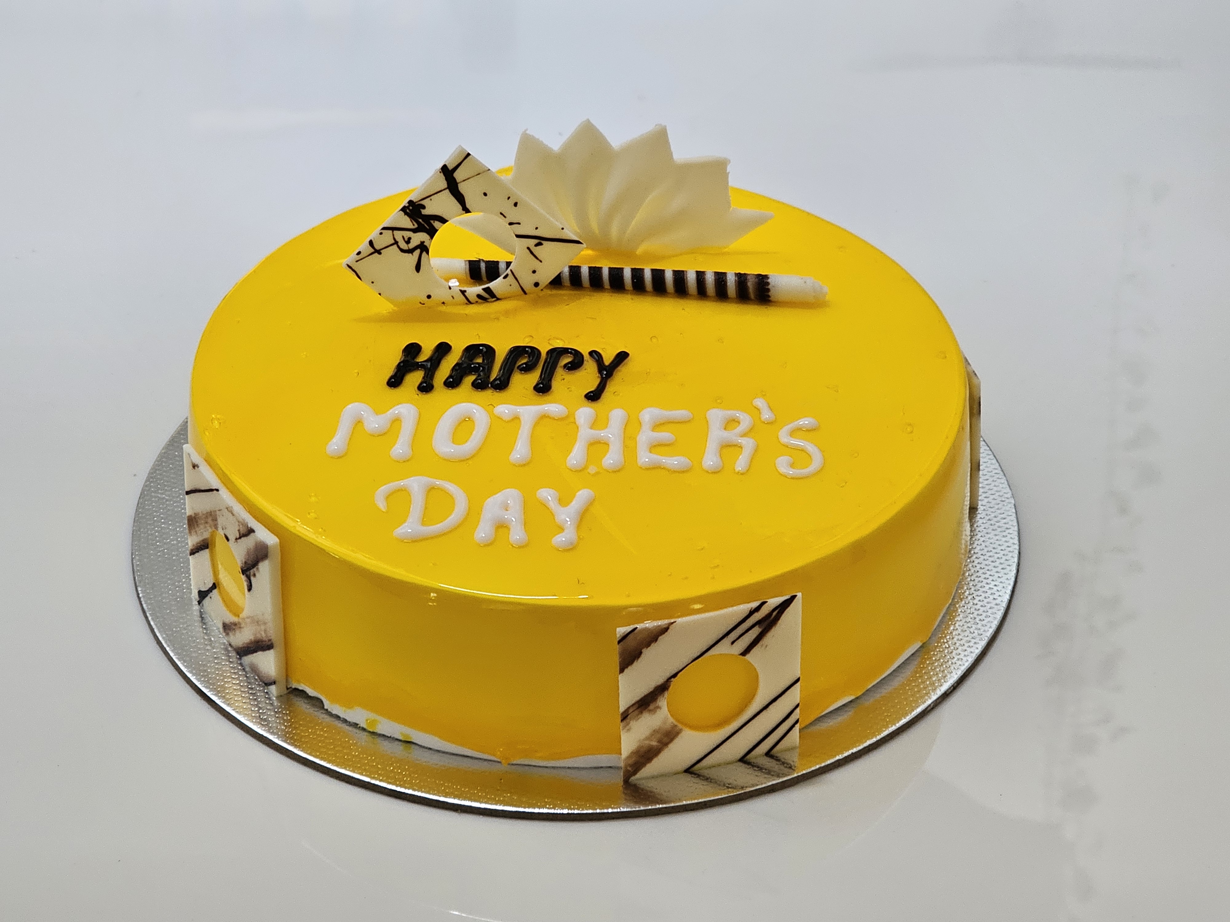 A round Mango cake with bright yellow frosting, decorated with white chocolate pieces and a white fondant flower. The cake has "HAPPY MOTHER'S DAY" written on it in white lettering and is placed on a silver cake board.