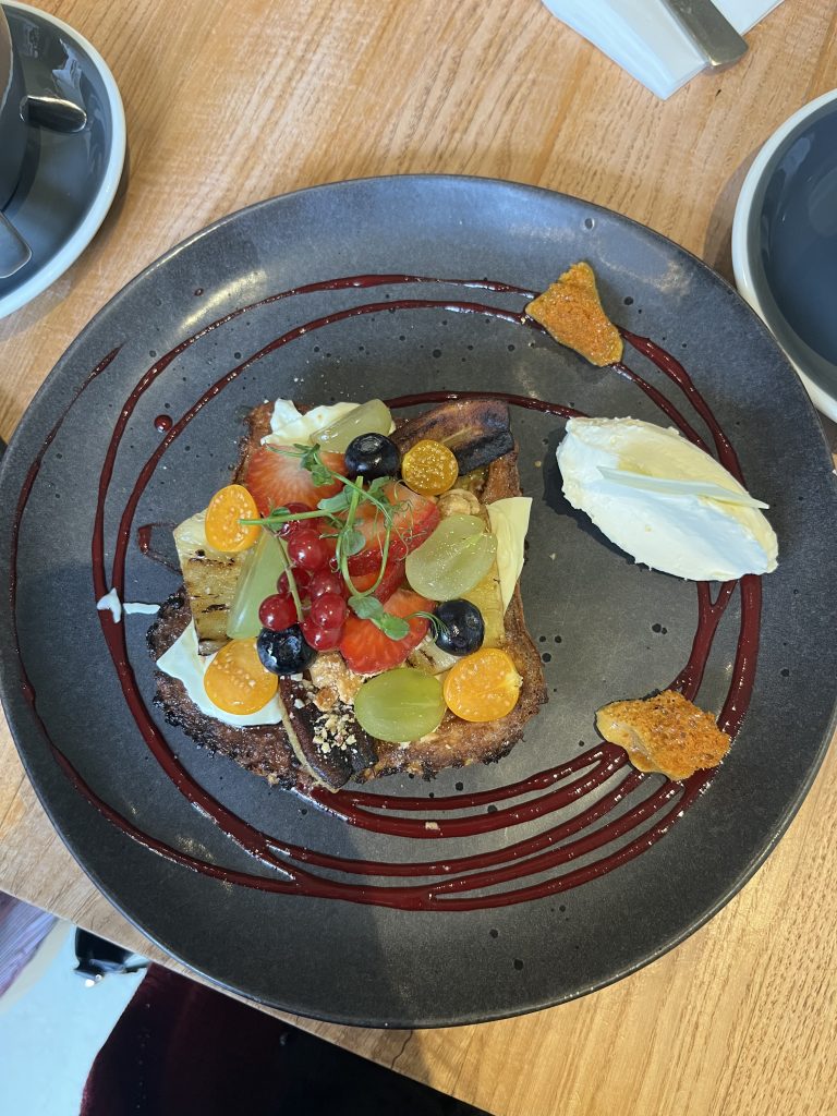 A gourmet dish served on a dark plate, featuring a piece of toast topped with assorted fresh fruits including strawberries, blueberries, grapes, red currants, and physalis. There are drizzles of a red sauce on the plate, with a side of whipped cream or butter and a crispy textured garnish.