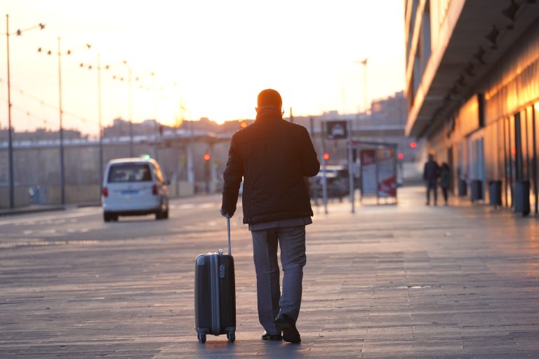 A person walking on a sidewalk with a wheeled suitcase in tow, approaching a distant taxi under a setting sun, adjacent to a train station.