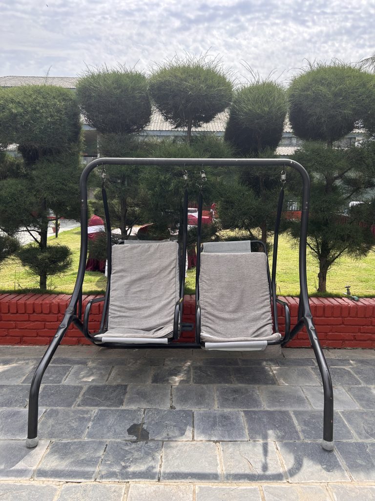 A metal-framed outdoor swing seat with two gray fabric cushions is positioned on a stone patio. In the background, there is a neatly trimmed row of green shrubs and a red brick border set against a clear sky.