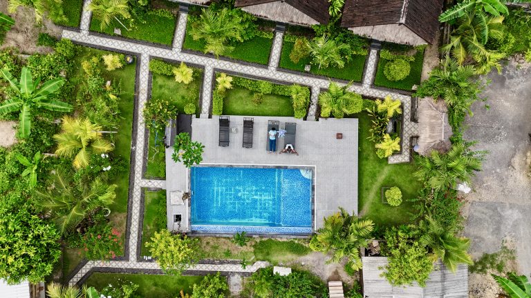 Aerial view of a tropical cottage with a swimming pool in the center, surrounded by lush greenery and palm trees. The pool area features lounge chairs and a paved deck, with two people relaxing by the poolside. Pathways with decorative tiles weave through the garden.