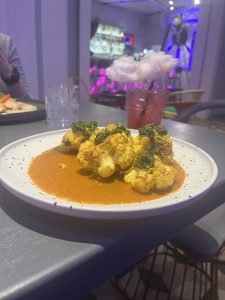 A plate of roasted cauliflower served with a red sauce and topped with green herbs. In the background, a drink with a straw and a cotton candy topper sits on the table, with colorful ambient lighting and bar shelves visible.