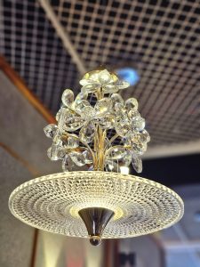  A crystal chandelier with flower-shaped glass elements and a textured glass shade hanging from a ceiling. The chandelier features a gold-toned frame and is illuminated, casting light and creating reflections on its various surfaces.  It was spotted in an electrical shop in Kozhikode, Kerala.
