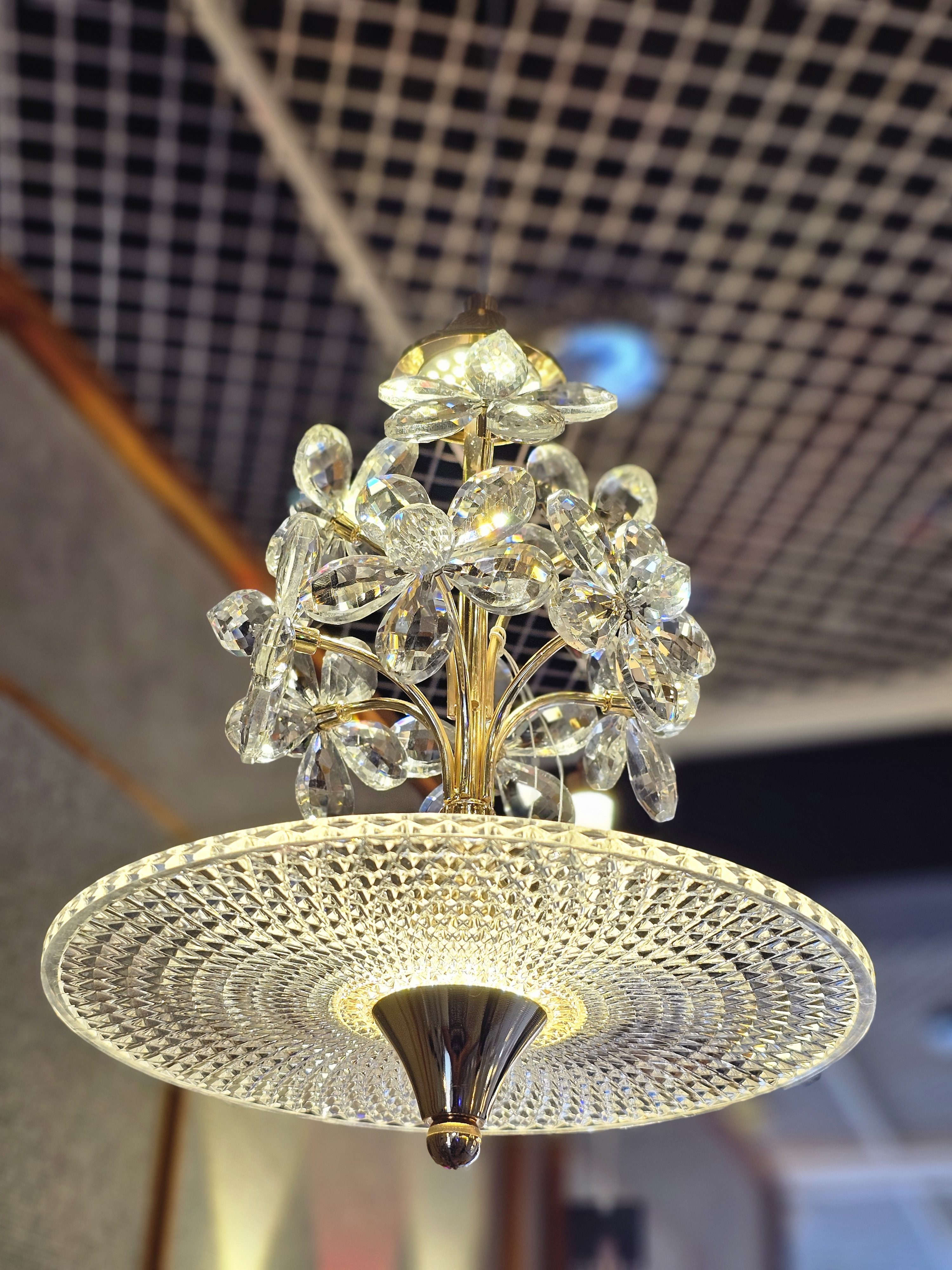 A crystal chandelier with flower-shaped glass elements and a textured glass shade hanging from a ceiling. The chandelier features a gold-toned frame and is illuminated, casting light and creating reflections on its various surfaces. It was spotted in an electrical shop in Kozhikode, Kerala.